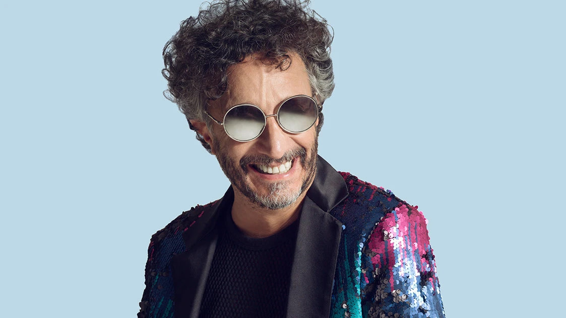 Fito Páez con anteojos redondos y saco de lentejuelas sobre fondo celeste